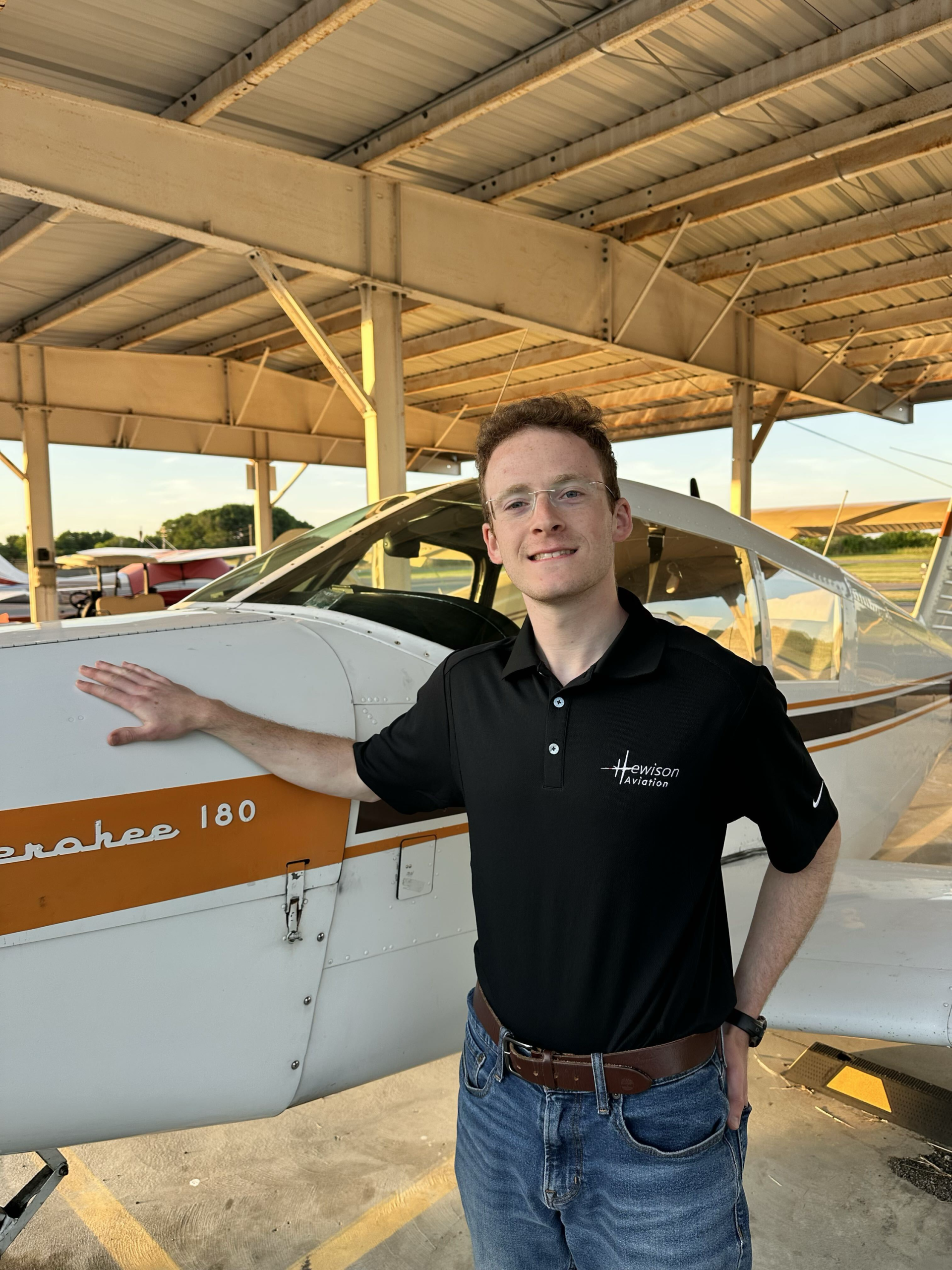 Smiling man in a black polo with a Hewison Aviation logo stands beside a small white and orange airplane in a hangar.