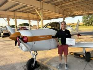 Man in black polo and red shorts stands beside a Cherokee 180 airplane in a hangar, holding a trophy and an award certificate.