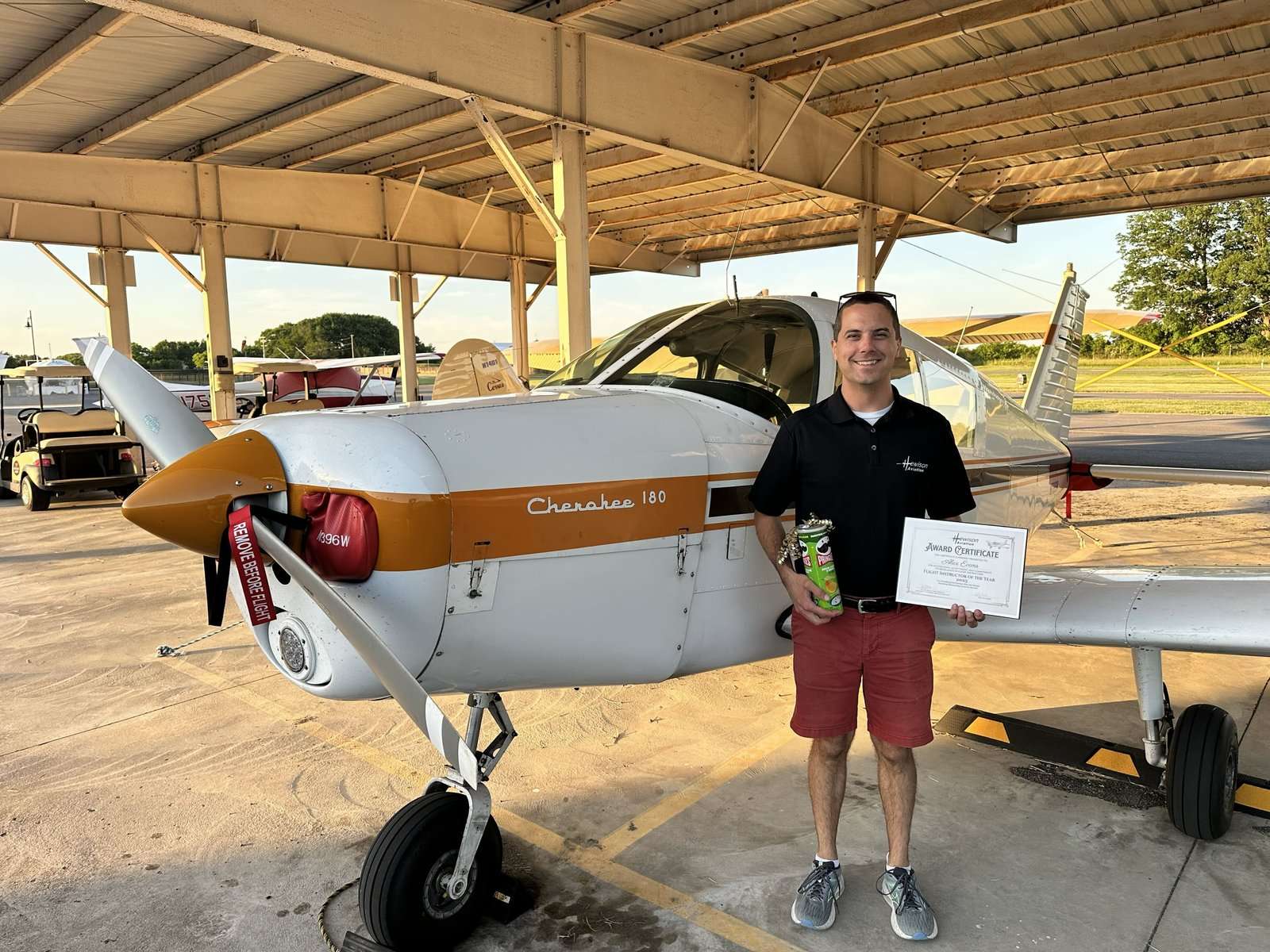 Man in black polo and red shorts stands beside a Cherokee 180 airplane in a hangar, holding a trophy and an award certificate.